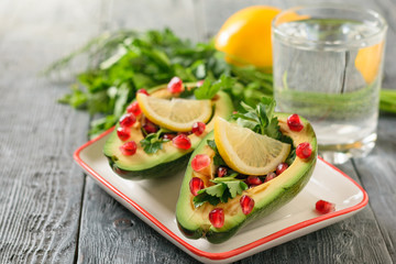 A glass of clean water with avocado halves filled with pomegranate seeds and parsley on a gray wooden table. Vegetarian cuisine for weight loss.