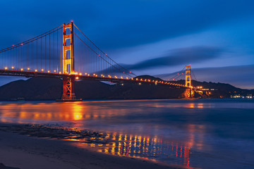 Nighttime view of Golden Gate Bridge reflected in the blurred water surface of San Francisco bay, dark blue sky background; California