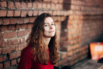 beautiful long-haired girl in a red dress is sitting on a brick wall background. The concept of a lonely woman.