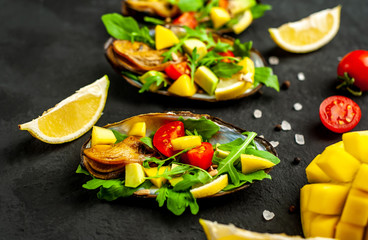 cooked mussels on a black concrete background with avocado, arugula, mango, tomatoes, lemon