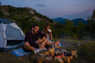 Naklejka premium Young couple relaxing by the campfire and drinking coffee in the forest hill in the dusk