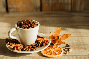 Cup with coffee beans and dried orange placed with star anise on wooden table lit by morning...