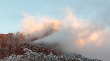 The first light of a winter morning gives the clouds shrouding Mt. Kinesava in Zion national park Utah a soft pink glow.