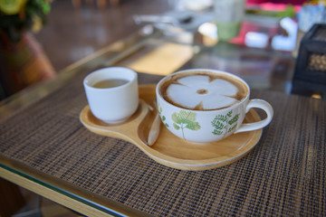 Latte coffee in a white cup, leaf pattern, paired with green tea in a small white cup placed on a wooden plate in a coffee shop.