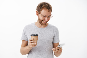 Morning starts with jokes in internet and coffee. Portrait of entertained happy carefree guy with bristle holding paper cup and smartphone smiling broadly at gadget screen typing message