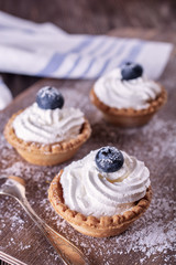 Bright tartlets filled with curd cream and decorated with blueberries on top are on a wooden desk and next to spoon, close up, vertical