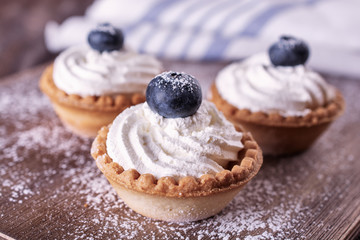 Bright tartlets filled with curd cream and decorated with blueberries on top are on a wooden desk and next to napkin, close up