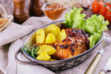 Appetizing meal with baked chicken leg and potato pieces with green lettuce leaves and rosemary in a ceramic plate on a linen napkin next to tomato sauce, spices, tomatoes and fork, close up