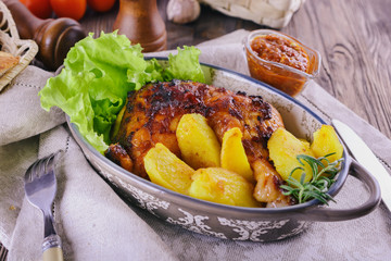 Appetizing meal with baked chicken leg and potato pieces with green lettuce leaves in a ceramic oval-shaped bowl on a linen napkin next to tomato sauce, spices and fork, close up