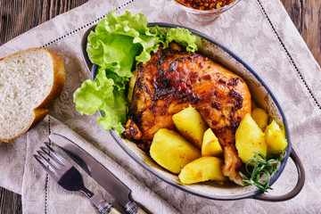Appetizing dish with baked chicken leg and potato pieces with green lettuce leaves in a ceramic oval-shaped bowl on a linen napkin next to fork and knife, top view