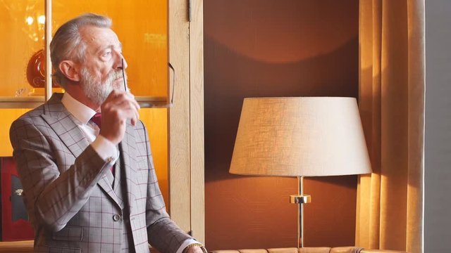 Portrait of whitebeard male with short grey haircut, dressed in tailored formal mens clothing posing with thoughtful look at luxury office interior, making serious business decision