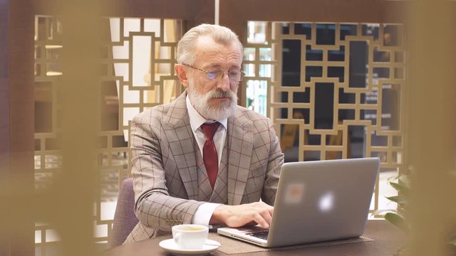 Eldely male successful scientist dressed in formal grey suit, working on laptop, searching important details of work, has confident serious expression, sitting at cafe table with cuo of coffee