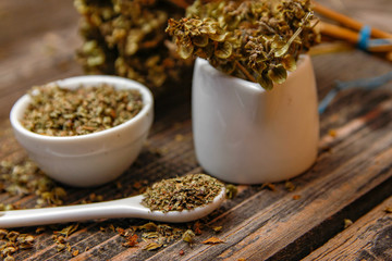 Dried basil spice in a small ceramic bowl, small white ceramic spoon and twig of dried basil on a rustic wooden table.