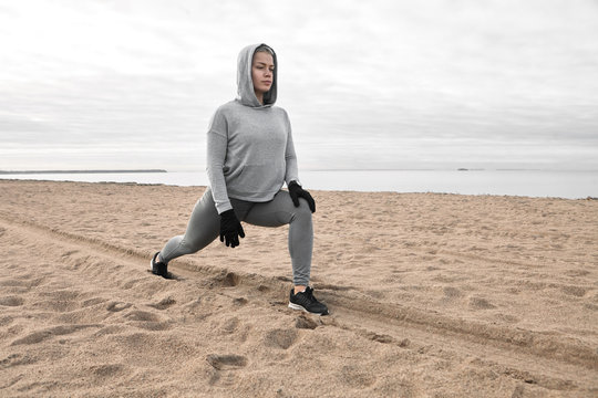 Isolated picture of athletic sporty girl in hoodie, leggings and sneakers warming up body, preparing leg muscles in crescent lunge pose on beach, having focused concentrated facial expression