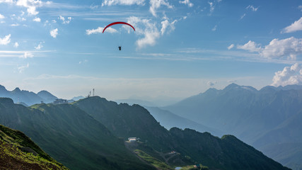 Red paraglider in blue cloudy sky over green mountains. Green valley with cable car down below. Krasnaya Polyana, Sochi, Caucasus, Russia.