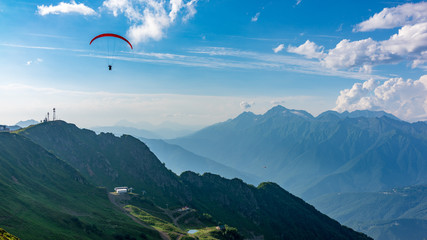 Fototapeta na wymiar Red paraglider in blue cloudy sky over green mountains. Green valley with cable car down below. Krasnaya Polyana, Sochi, Caucasus, Russia.