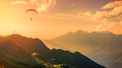 Red paraglider in the orange sunset cloudy sky over the green mountains. Green valley with cable...