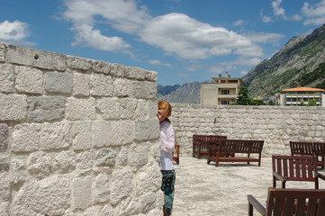 Fototapeta premium Red-haired girl looks out from behind a stone wall on a sunlit terrace