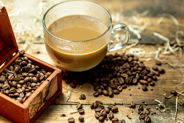 A glass cup of coffee with milk in the morning, next to roasted coffee beans, on a wooden, natural, rustic table
