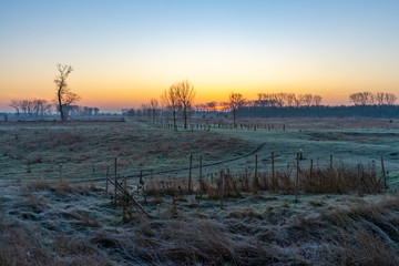 A winter scene with a path through a Dutch river delta landscape on the Belgian border. The rising sun colors the sky orange and blue.