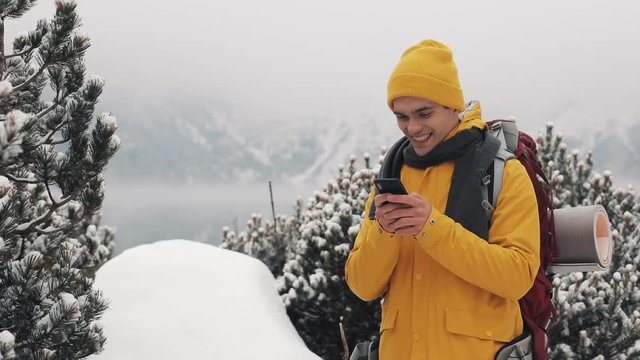 Young traveler man using smartphone in hiking winter tour. Snowy blurred mountains cape and lake. Traveling and communication concept: hiker scrolling and tapping on line by cell phone