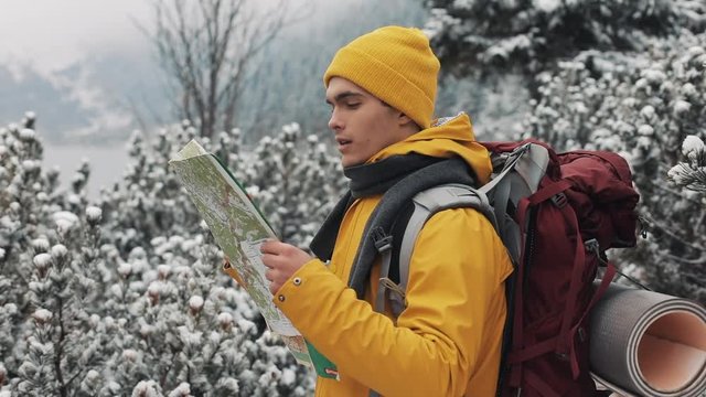 Traveling in the mountains. Young man wearing yellow winter clothing holding map standing in the woods covered with snow. Trip, adventure, climbing