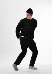 Young dancer dressed in black jeans, sweatshirt, hat and gray sneakers is dancing freestyle in the studio on the white background