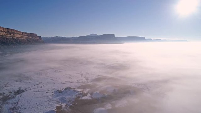 Panning aerial view of thin layer of fog over snow in the desert as the sun shines high in the blues sky.