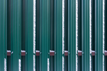 The fence is made of metal plates of green color with adhering snow.