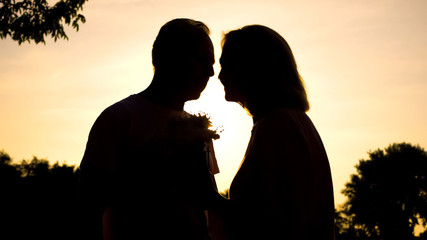 Aged married couple kissing in twilight park, holding flowers, romantic gift