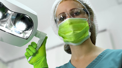 Lady dentist in protective uniform adjusting light before examination, closeup - Powered by Adobe
