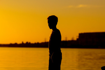 Fototapeta premium Silhouette of a boy standing near the river at sunset. Beach and calm water by the lake or river in the evening.