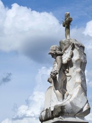 Scene in a cemetery: old stone statue of an angel holding a bunch of flowers. Above, a cross. In the background, a sky with some white clouds. Sunny day.