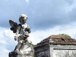 Scene in a graveyard: old stone statue with wings. In the background, a cloudy sky. Sunny day.