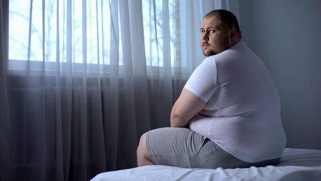 Sad fat man sitting on bed at home, looking at camera, depression, insecurities