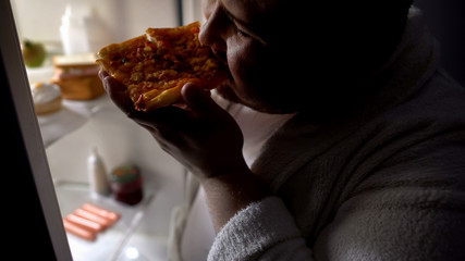 Unmotivated obese bachelor eating pizza near fridge at night, diet failure