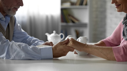 Senior male holding his wifes hand, sitting at table with cup of tea, happiness