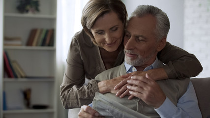 Elderly female giving hug for husband sitting on couch and reading newspaper