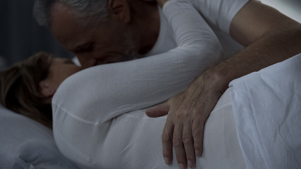 Gentle aged man kissing and hugging woman, husband and wife lying in bed