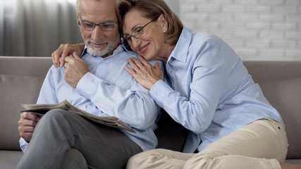 Happy retiree couple sitting on couch, man reading newspaper, lady hugging him