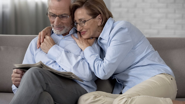 Happy aged man and lady sitting on couch, man reading newspaper wife hugging him
