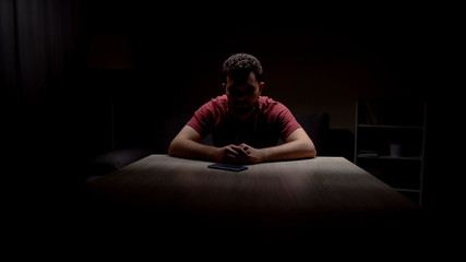 Depressed single man sitting in dark room looking at phone, waiting for call - obrazy, fototapety, plakaty