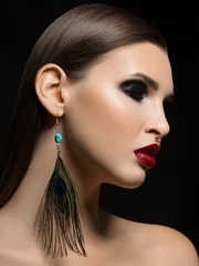 A close-up portrait of the beauty of a ryunette woman with gorgeous face features, which emphasizes the fatal make-up of red lipstick and fashionable peacock feather earring adds glamor to the image