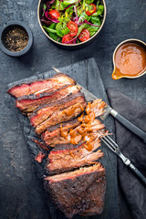 Traditionelles smoked barbecue Wagyu Beef Brisket angeboten als Draufsicht  auf einem alten verkohlten Board mit Louisiana Sauce und Salat mit Tomaten