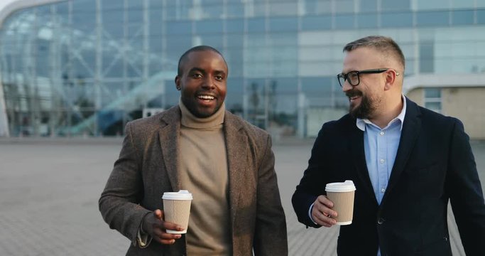 Portrait shot of the two attractive multiethnical men in business style walking outdoor at the big modern glass building with cofee in hands and speaking joyfully.