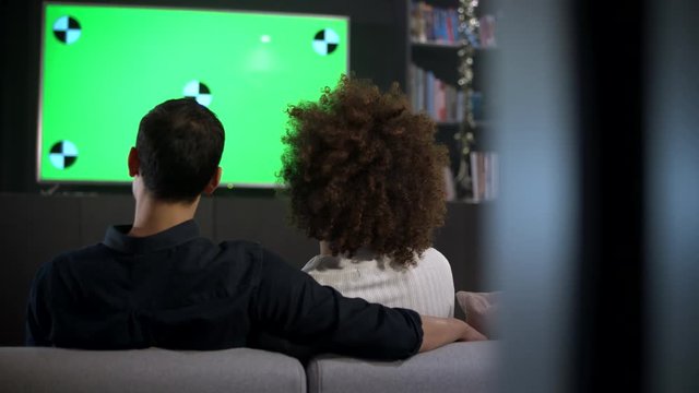 View From Behind A Young Couple Sitting On A Sofa Watching TV Together. Track Left And Right.