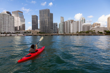 Adventurous girl kayaking in front of a modern Downtown Cityscape during a sunny evening. Taken in Miami, Florida, United States of America.