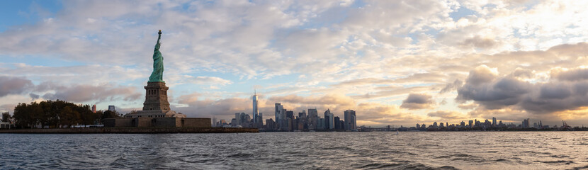 Fototapeta premium Panoramic view of the Statue of Liberty and Downtown Manhattan in the background during a vibrant cloudy sunrise. Taken in Jersey City, New Jersey, United States.