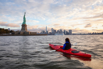 Adventurous Girl Sea Kayaking near the Statue of Liberty during a vibrant cloudy sunrise. Taken in Jersey City, New Jersey, United States.