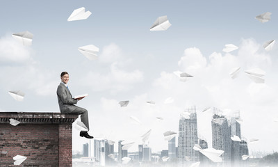 Man on roof edge reading book and cityscape at background